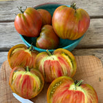 Load image into Gallery viewer, A collection of 'Taiga' tomatoes with a green, bronze, and red streaked rind, cut open to reveal the marbled interior, on a wooden surface with a knife and a blue bowl in the background.
