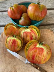 Load image into Gallery viewer, A collection of 'Taiga' tomatoes with a green, bronze, and red streaked rind, cut open to reveal the marbled interior, on a wooden surface with a knife and a blue bowl in the background.
